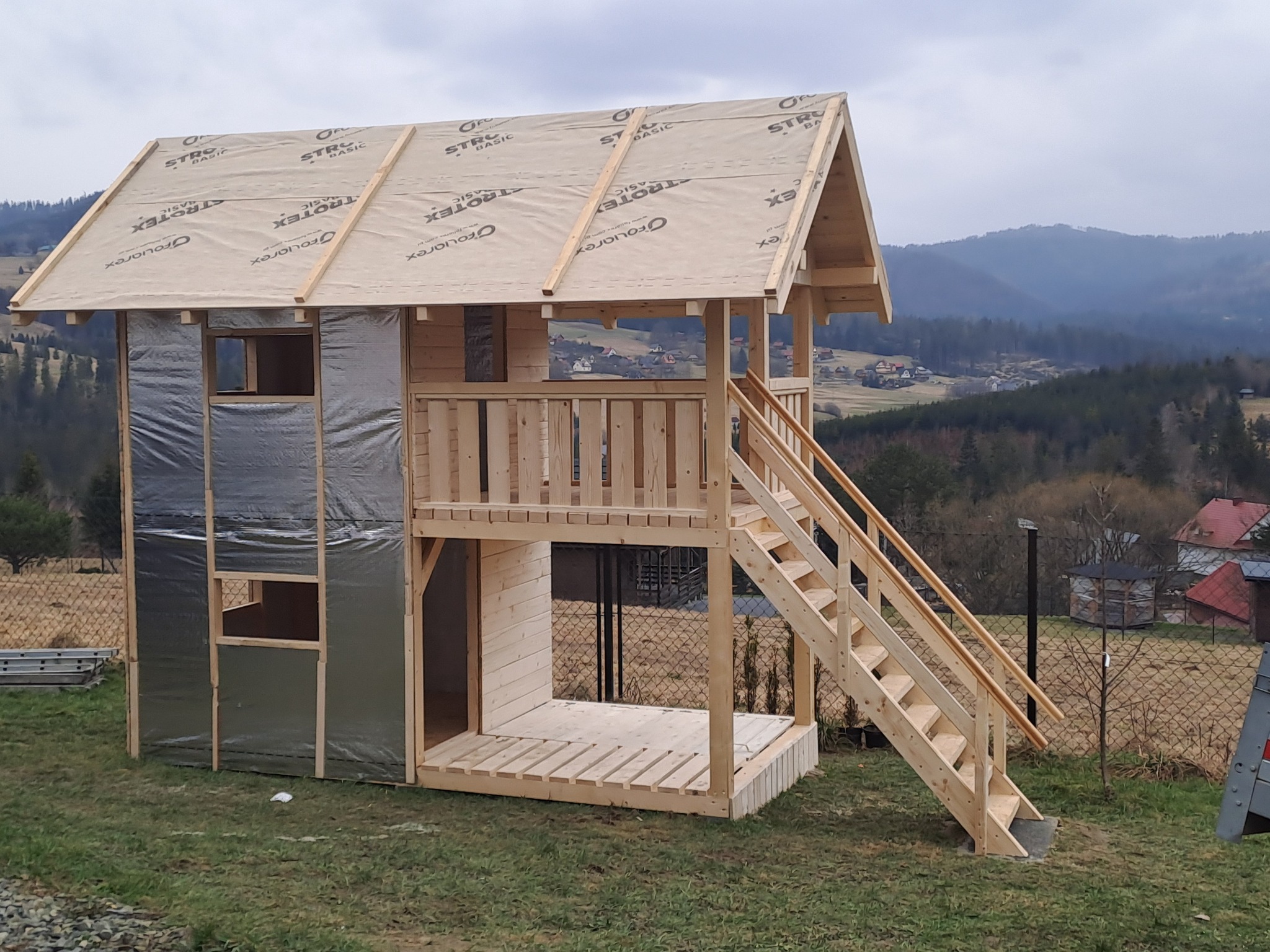 Dwukondygnacyjna konstrukcja z surowego drewna z zadaszeniem pokrytym membraną, prawdopodobnie plac zabaw dla dzieci, z widocznymi elementami izolacji na ścianach, umieszczona na trawiastym terenie...