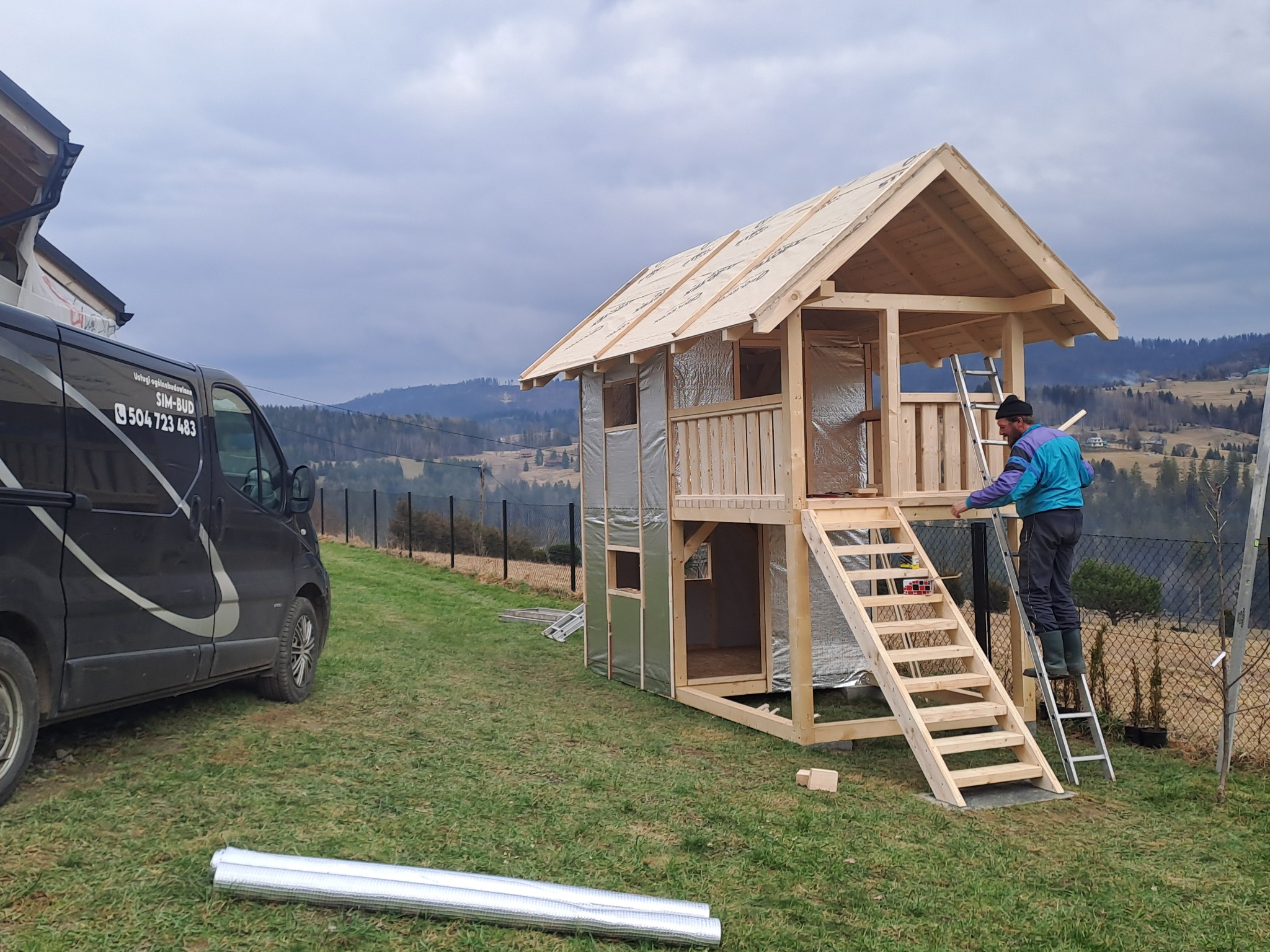 Budowa dwupoziomowego domku z drewna z izolacją, pracownik na drabinie, w tle górzysty krajobraz i samochód firmy budowlanej.
