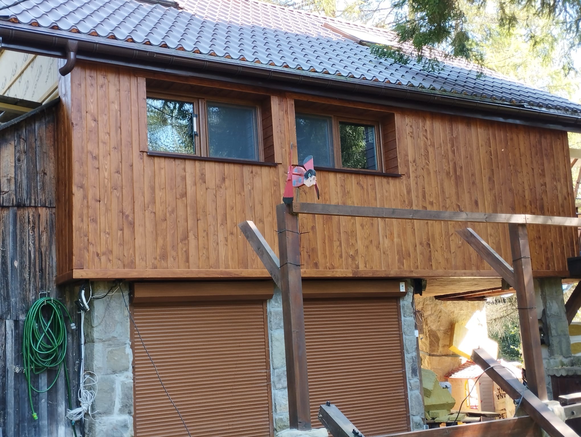 Dwukondygnacyjny budynek z drewnianą elewacją i brązową dachówką, z roletami w oknach garażowych na kamiennej podstawie, ozdobiony wiatraczkiem na słupku.