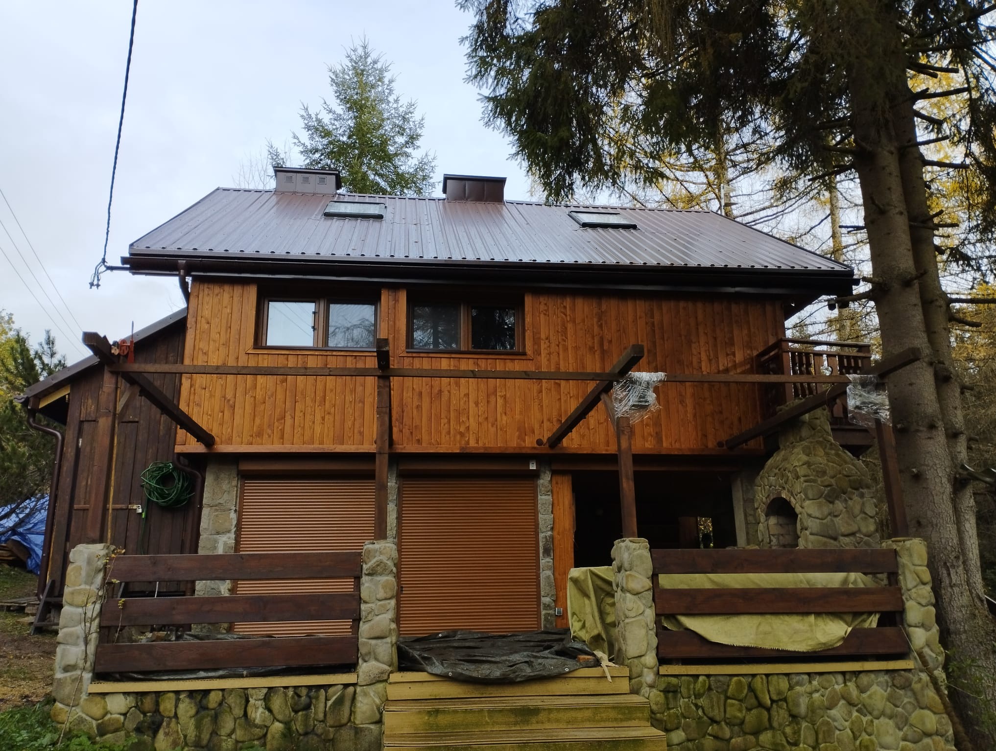 Dwukondygnacyjny dom z drewnianą elewacją i brązowym, metalowym dachem. Kamienna podmurówka i elementy dekoracyjne z kamienia. Częściowo zasłonięte rolety zewnętrzne.