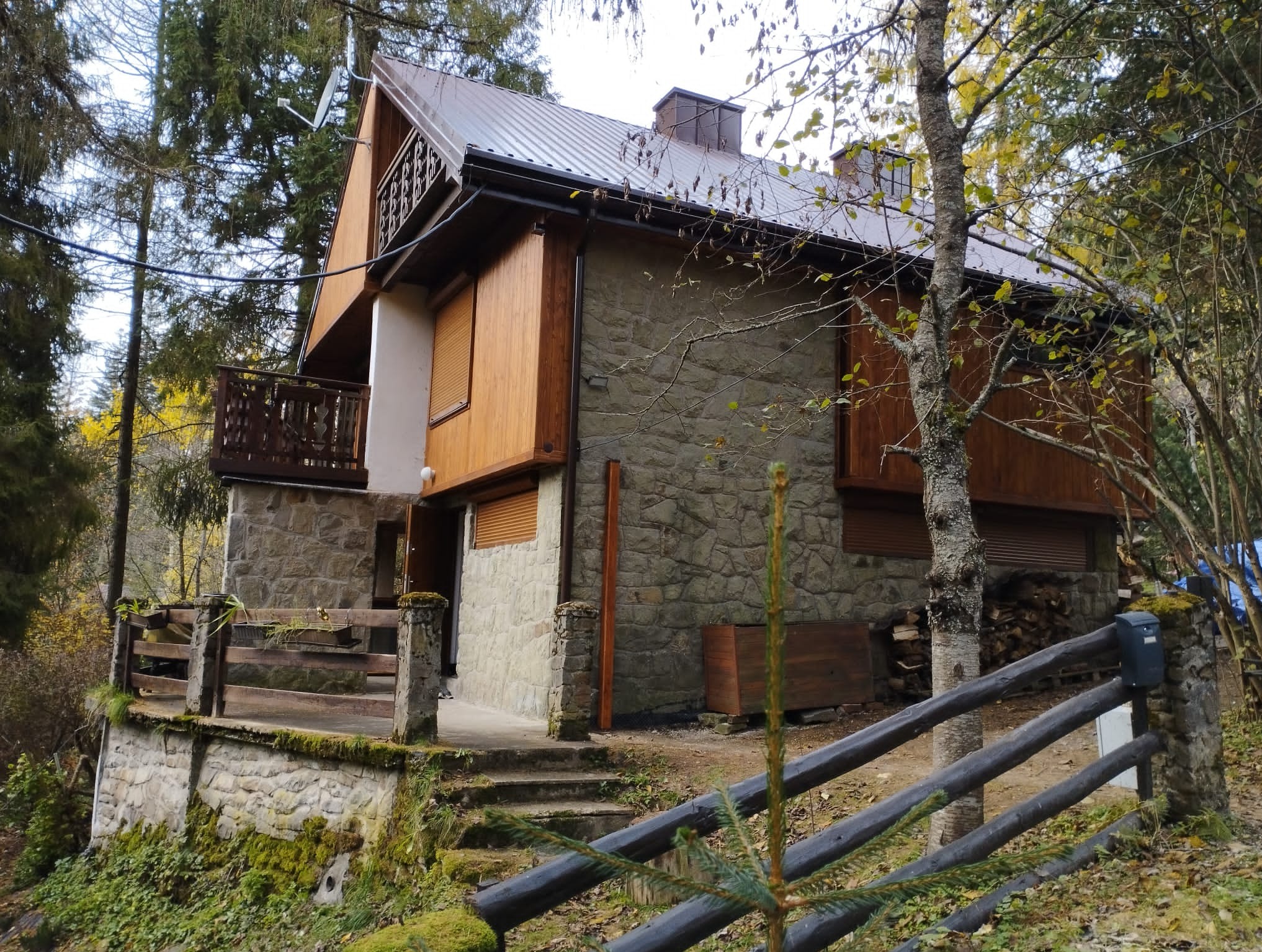 Dwukondygnacyjny dom z kamienną elewacją i drewnianymi elementami, otoczony lasem, widoczny z zewnątrz, z metalowym dachem i drewnianym balkonem.