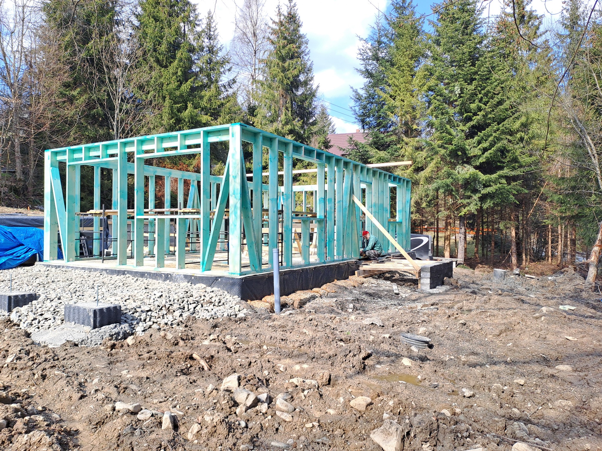 Wznoszenie konstrukcji szkieletowej domu na fundamencie, widoczne impregnowane drewniane elementy szkieletu, gruz i fundamenty, w tle las iglasty.