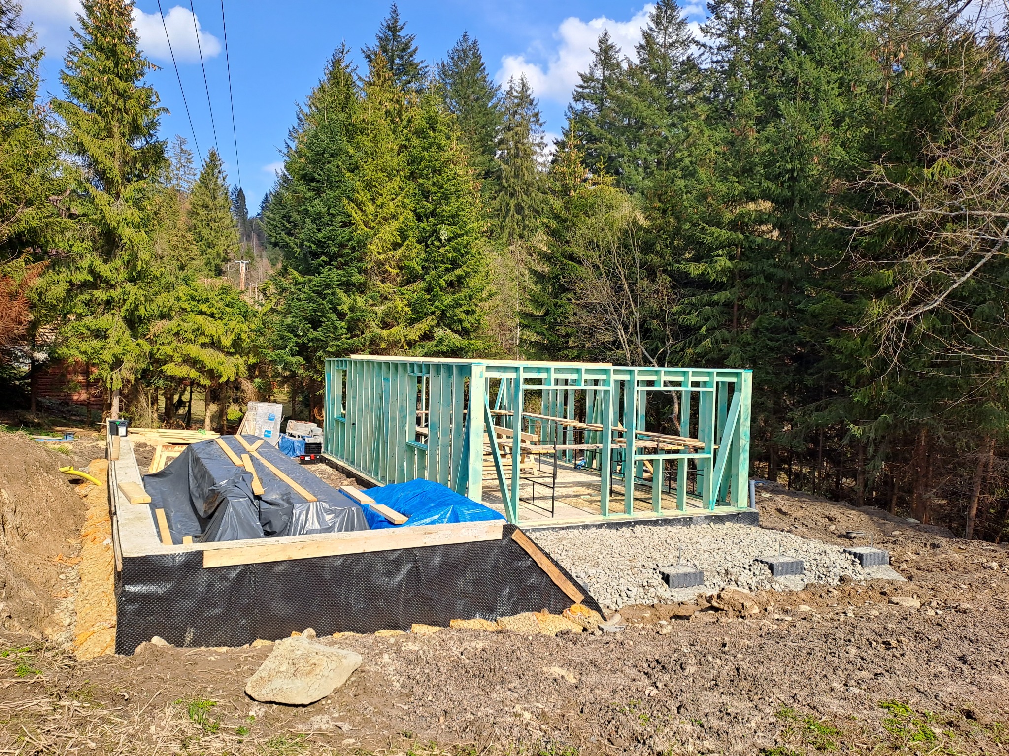 Wznoszenie szkieletu domu na przygotowanym fundamencie z hydroizolacją, w tle las iglasty i linia energetyczna.