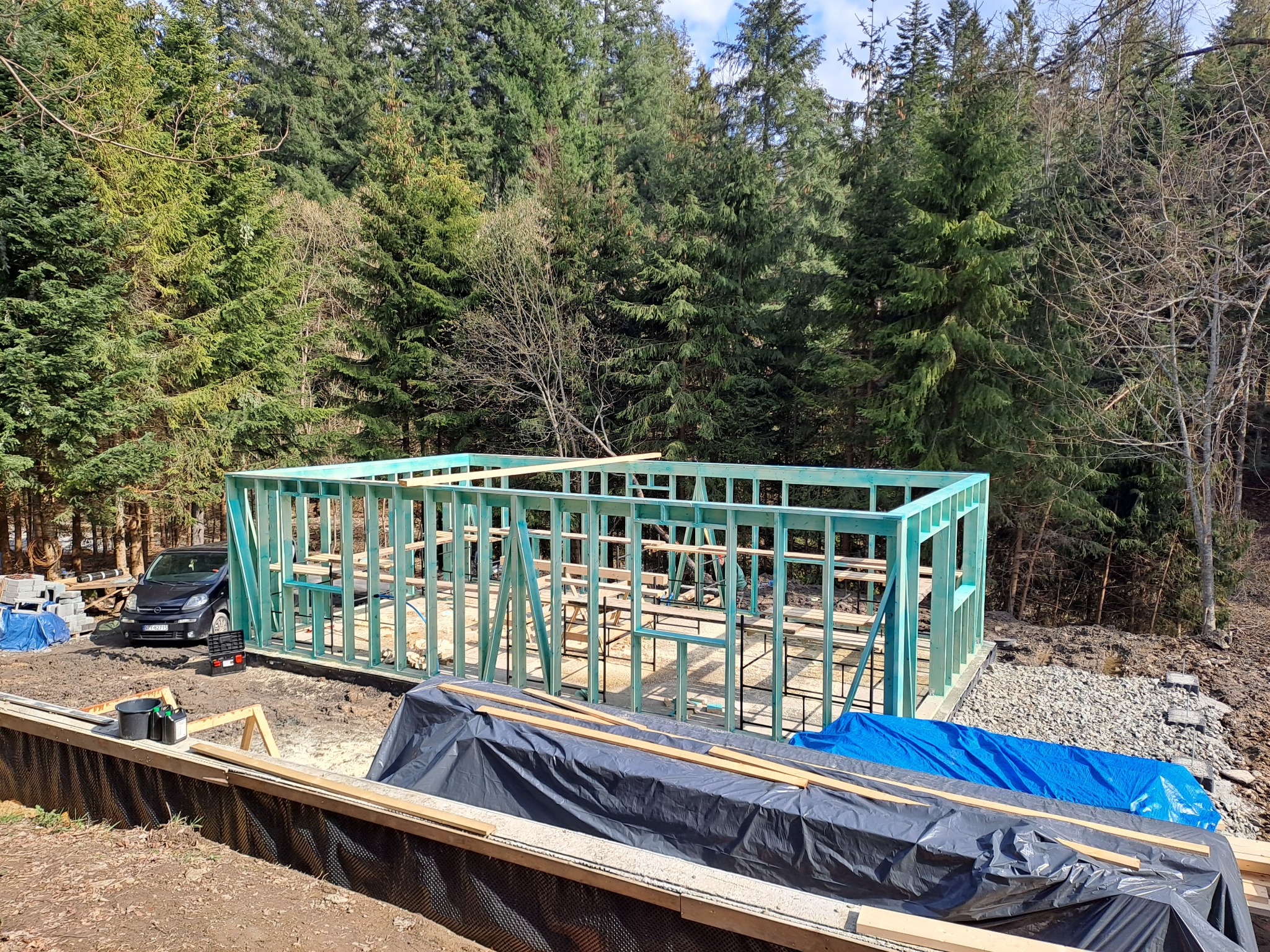 Wznoszenie szkieletu domu na fundamencie, widoczne drewniane elementy konstrukcyjne pomalowane na zielono, w tle las i zaparkowany samochód.