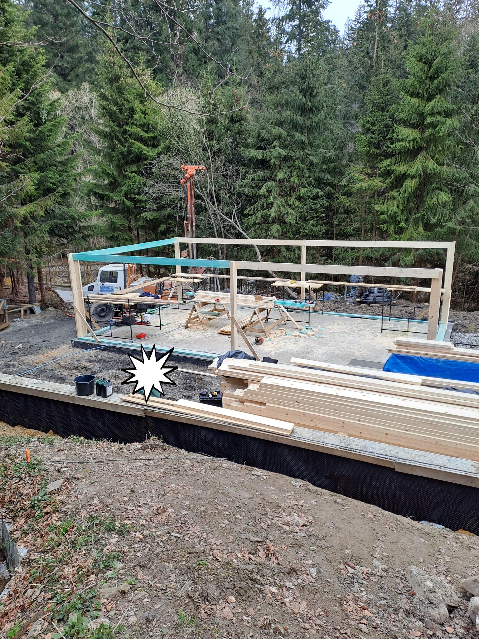 Budowa domu w lesie: widok na szkielet konstrukcji z belek, przygotowane deski, fundament zabezpieczony folią kubełkową, w tle las i dźwig