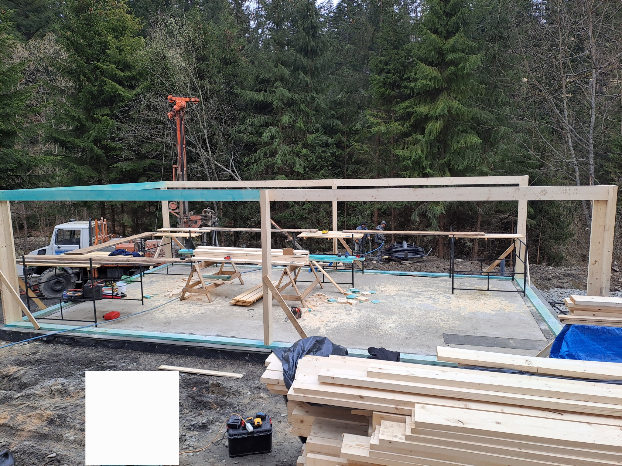 Wznoszenie drewnianej konstrukcji na betonowej płycie fundamentowej w otoczeniu drzew; widoczny sprzęt budowlany i narzędzia, w tle wiertnica.