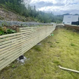 Montaż drewnianego ogrodzenia panelowego na nierównym terenie, widoczne betonowe podstawy słupków i częściowo ułożone panele, w tle zbocze porośnięte zielenią i nowoczesny budynek.