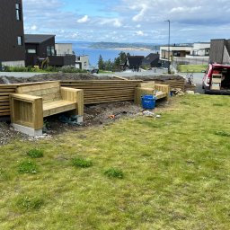 iForm - Dobrze Zbudowany s.p. Z.o.o. - Budowa drewnianej konstrukcji z siedziskami w ogrodzie, widoczny samochód dostawczy z narzędziami, w tle zabudowa mieszkalna i widok na morze.