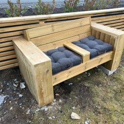 iForm - Dobrze Zbudowany s.p. Z.o.o. - Drewniana ławka z szarymi poduszkami w ogrodzie, obok drewnianego płotu, z widoczną ziemią i trawą.