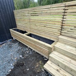 iForm - Dobrze Zbudowany s.p. Z.o.o. - Drewniana konstrukcja z poziomych listew tworząca ściankę i donicę, w tle zielona roślinność, na ziemi szary żwir.