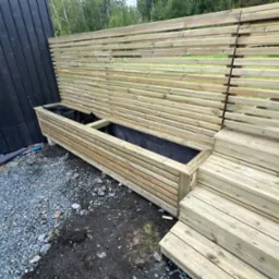 Drewniana konstrukcja z poziomych listew tworząca ściankę i donicę, w tle zielona roślinność, na ziemi szary żwir.