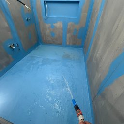 iForm - Dobrze Zbudowany s.p. Z.o.o. - Hydroizolacja w narożnej kabinie prysznicowej z wnęką, ściany pokryte szarą farbą z niebieskimi akcentami, woda spryskiwana z węża w celu sprawdzenia szczelności.