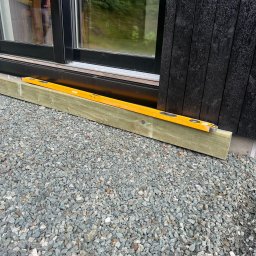 iForm - Dobrze Zbudowany s.p. Z.o.o. - Poziomica spoczywa na drewnianej belce przy czarnej fasadzie budynku z widocznym oknem i żwirowym podłożem.