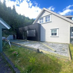 iForm - Dobrze Zbudowany s.p. Z.o.o. - Przygotowanie podłoża pod taras z drewnianą ramą na żwirowej podsypce, widok na dom z białą elewacją i czarną dobudówką, w tle zalesione wzgórze.