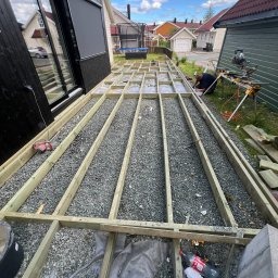 iForm - Dobrze Zbudowany s.p. Z.o.o. - Konstrukcja drewnianego tarasu na żwirowym podłożu w trakcie budowy, widoczny pracownik przy pracy oraz narzędzia i materiały budowlane.