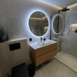 Nowoczesna łazienka z okrągłym lustrem podświetlanym LED, drewnianą szafką pod umywalką, czarną toaletą i kabiną prysznicową typu walk-in.