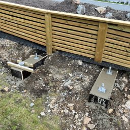 iForm - Dobrze Zbudowany s.p. Z.o.o. - Montaż drewnianego ogrodzenia panelowego: betonowe fundamenty z metalowymi kotwami w ziemi, widoczna perforowana folia izolacyjna przy panelach.