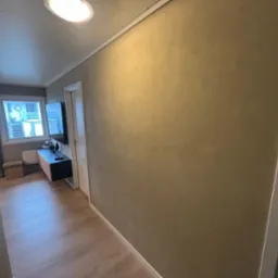 Beżowa ściana z efektem dekoracyjnym w przedpokoju, widok na drewnianą podłogę i okno z widokiem na budynek.