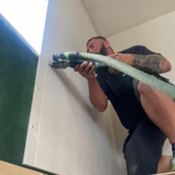 Mężczyzna z tatuażami docina płytę kartonowo-gipsową przy użyciu narzędzia wielofunkcyjnego Festool z podłączonym odkurzaczem, widoczna zielona ściana w tle.