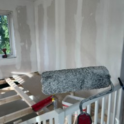 iForm - Dobrze Zbudowany s.p. Z.o.o. - Wałek malarski z resztkami białej farby na tle surowych ścian z nałożoną gładzią, widoczny fragment drewnianej konstrukcji podłogi i schodów w jasnym pomieszczeniu z oknem.