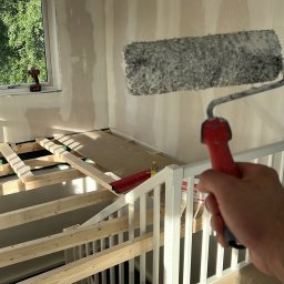 iForm - Dobrze Zbudowany s.p. Z.o.o. - Malowanie ścian wałkiem w trakcie remontu, widoczne drewniane elementy konstrukcyjne, zabezpieczona podłoga folią, ściany częściowo pokryte gładzią, w tle okno z zielenią.