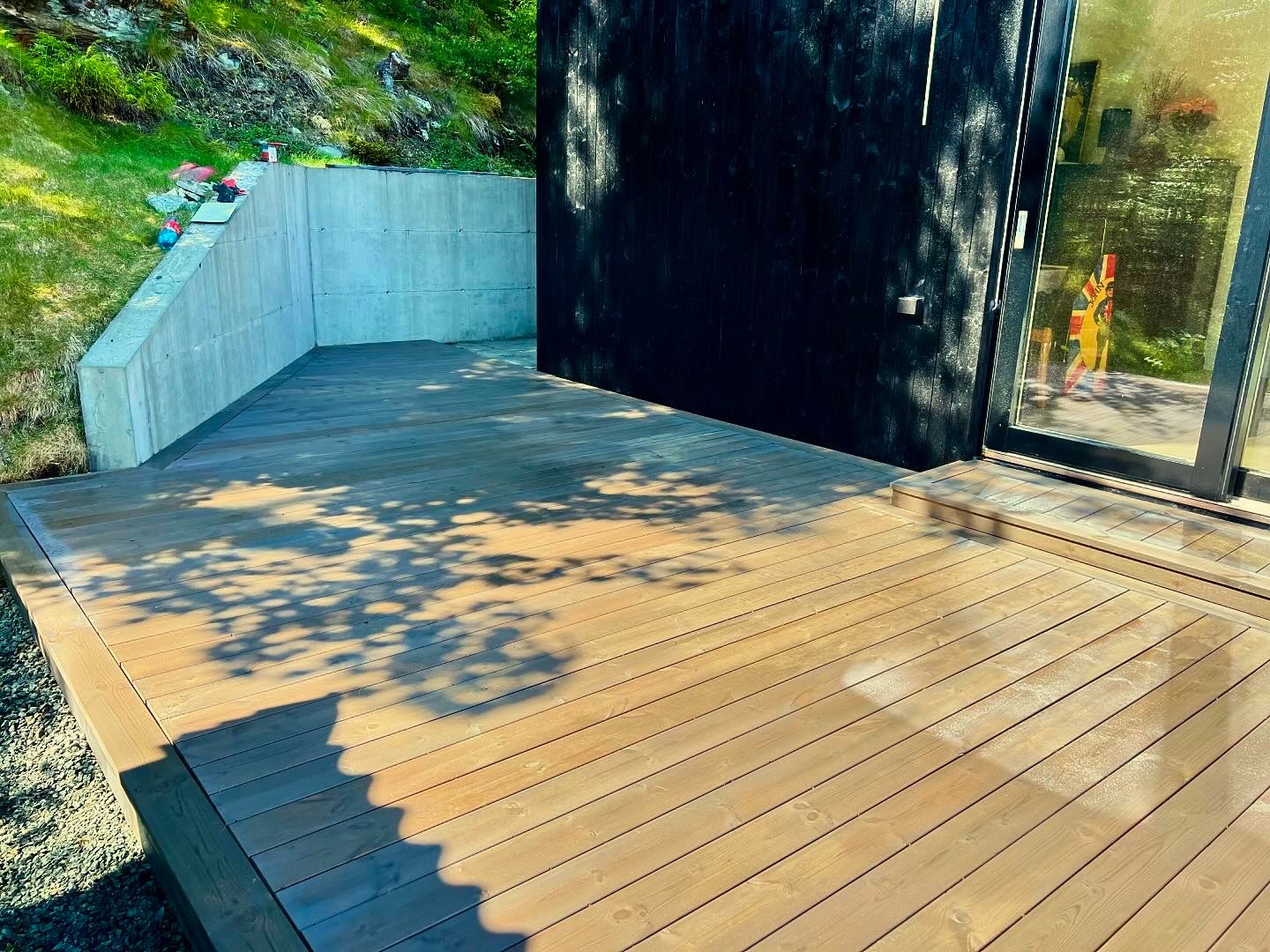 Nowoczesny taras z desek kompozytowych w odcieniach brązu, przylegający do czarnej elewacji budynku z dużym przesuwnym oknem, w tle betonowy murek oporowy i zielony trawnik z drzewami.