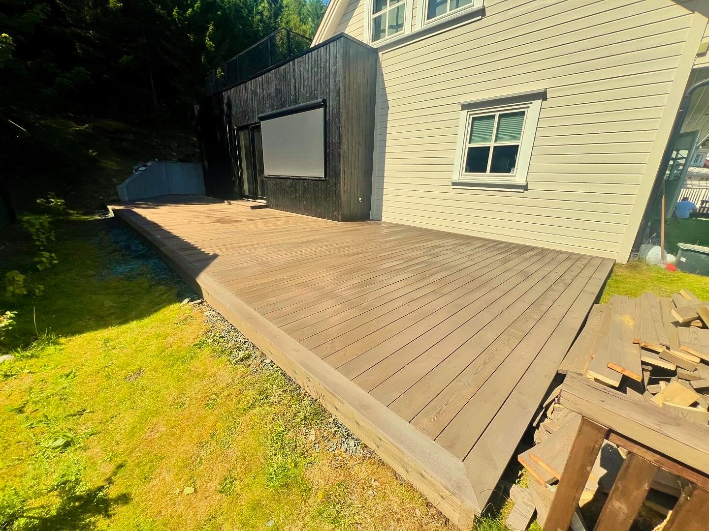 Nowo wybudowany, drewniany taras przylegający do domu z jasną elewacją, z widocznymi deskami na trawie, sugerującymi trwające prace wykończeniowe.
