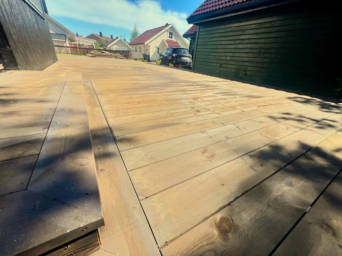 Nowo wybudowany, jasny drewniany taras zewnętrzny z widocznymi słojami drewna, prowadzący do domu z czerwonym dachem, w tle zaparkowany czarny samochód.
