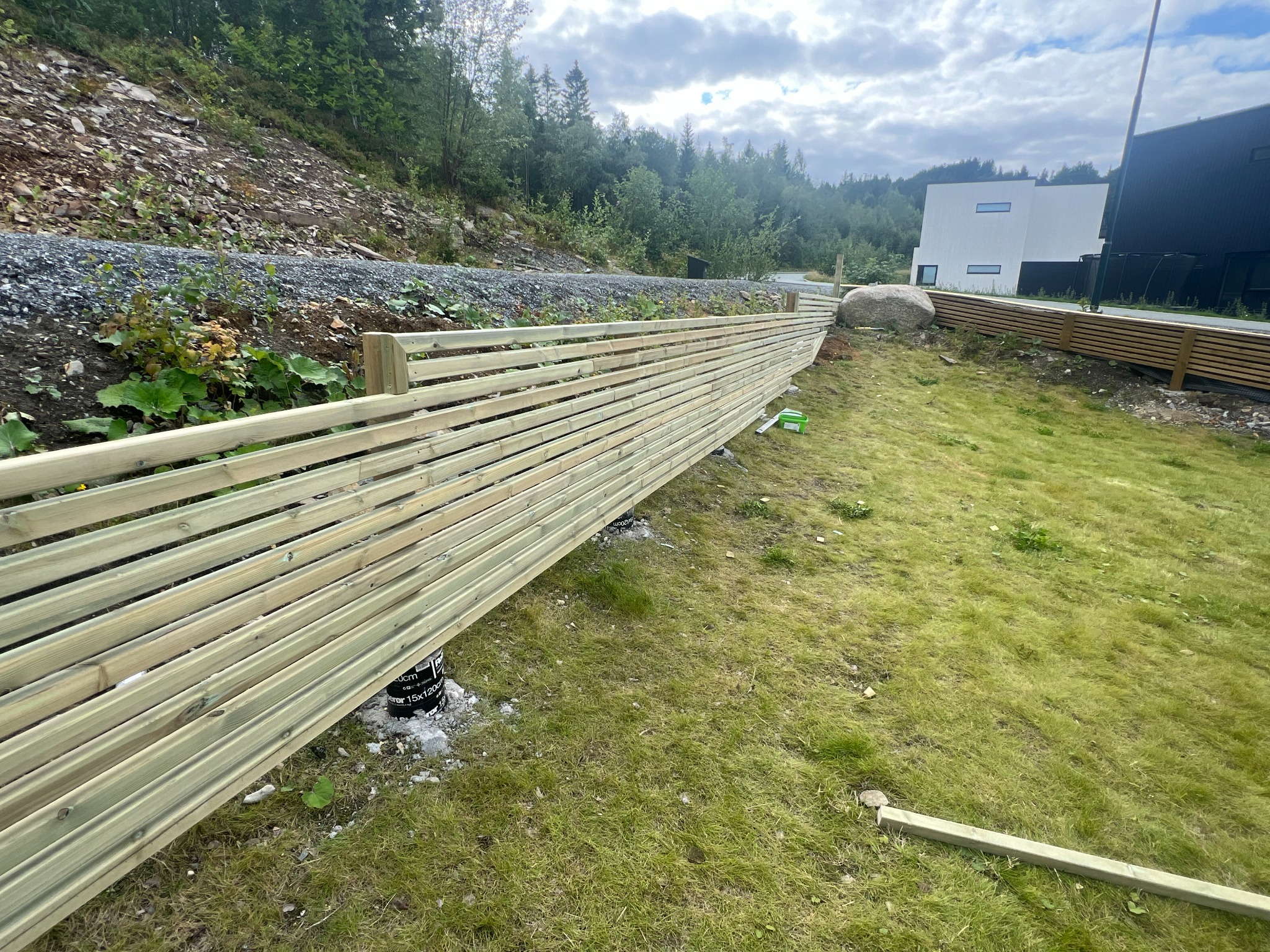 Montaż drewnianego ogrodzenia panelowego na nierównym terenie, widoczne betonowe podstawy słupków i częściowo ułożone panele, w tle zbocze porośnięte zielenią i nowoczesny budynek.