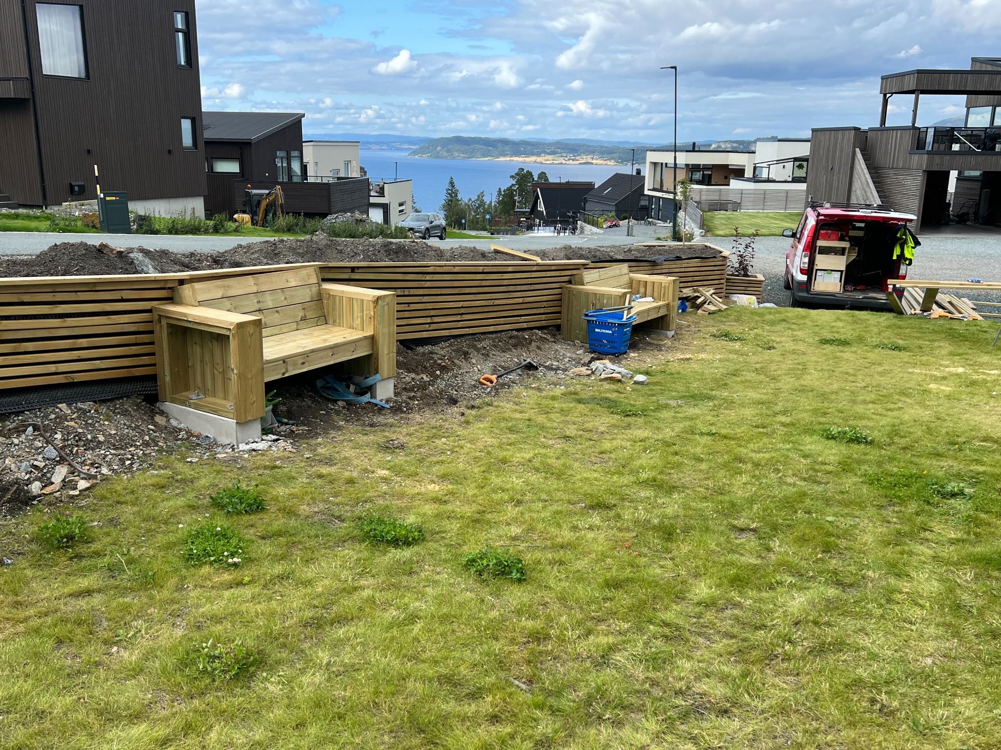 Budowa drewnianej konstrukcji z siedziskami w ogrodzie, widoczny samochód dostawczy z narzędziami, w tle zabudowa mieszkalna i widok na morze.