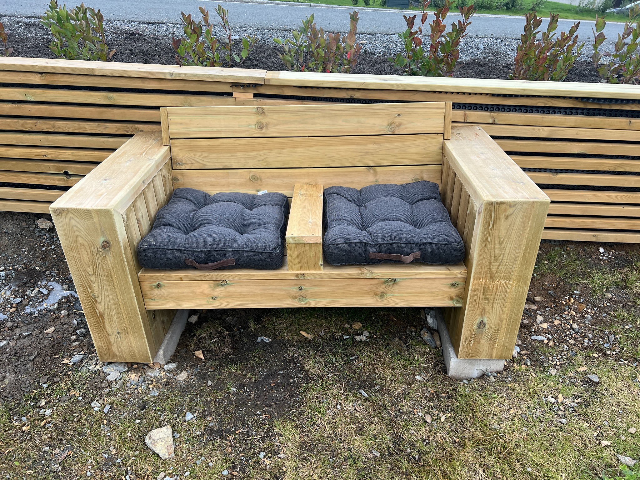 Nowoczesna ławka ogrodowa z jasnego drewna z dwoma szarymi poduszkami na siedzisku, ustawiona na betonowych podstawach na trawniku.