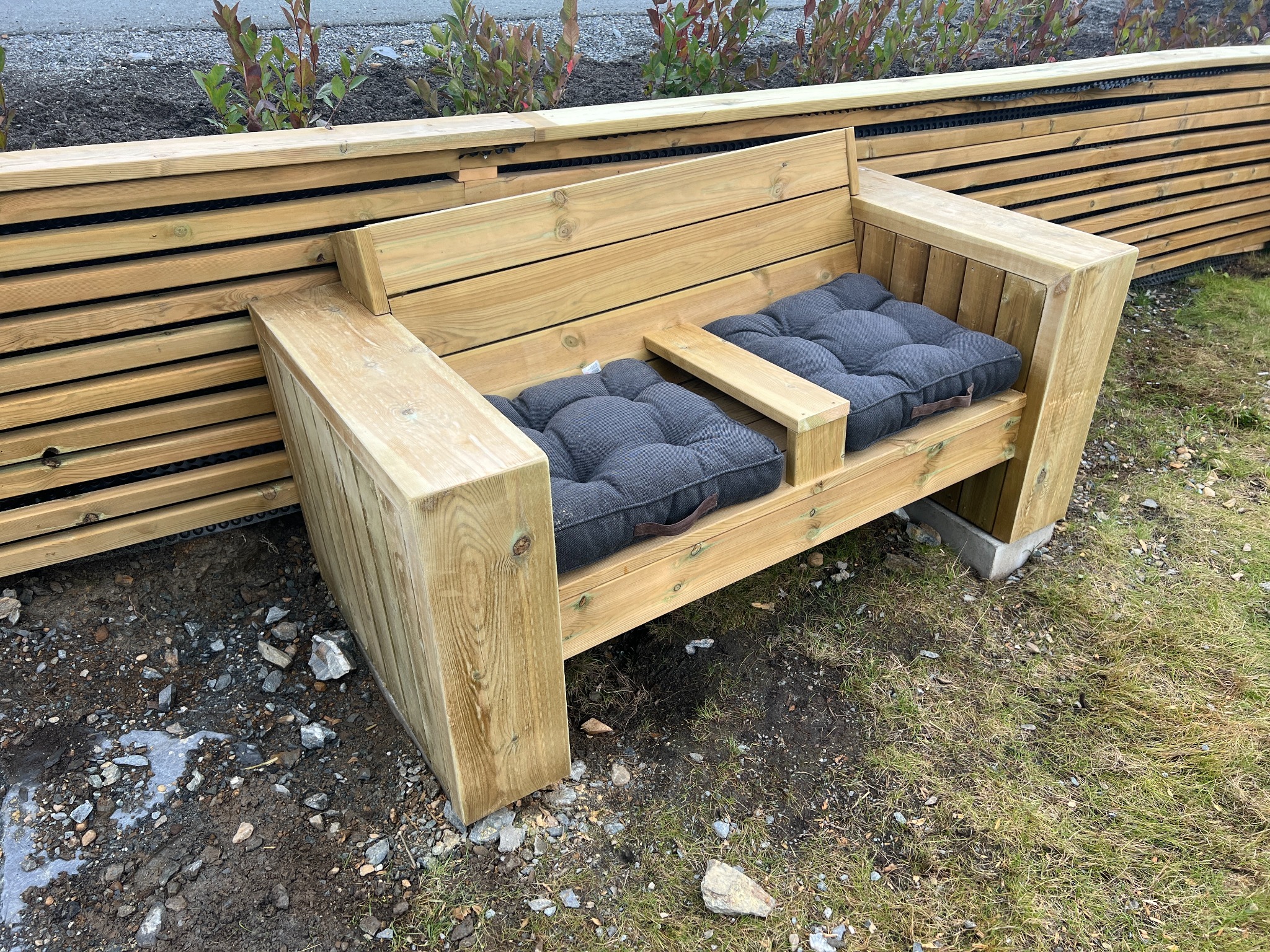 Drewniana ławka z szarymi poduszkami w ogrodzie, obok drewnianego płotu, z widoczną ziemią i trawą.