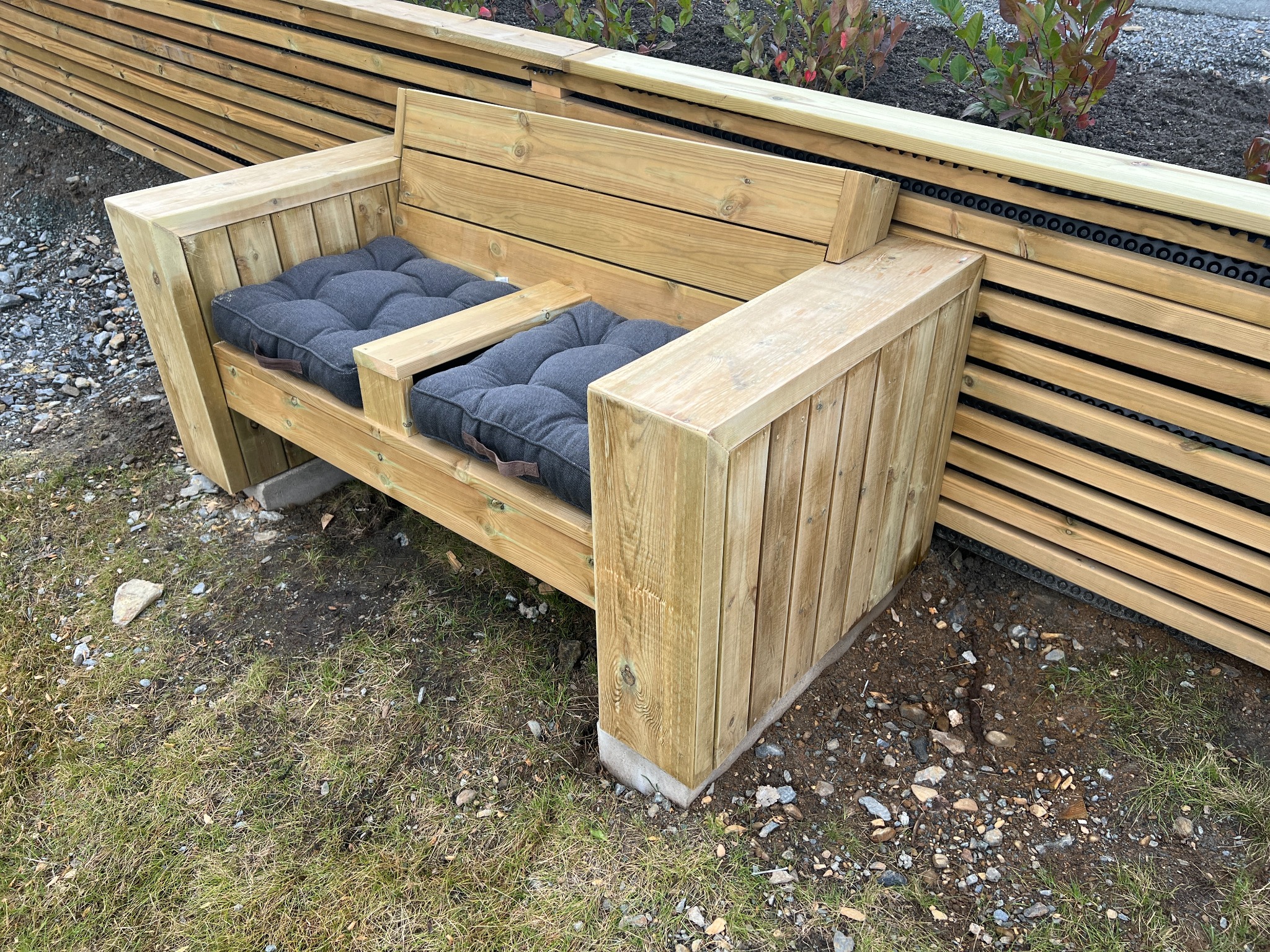 Nowoczesna drewniana ławka ogrodowa z szarymi poduszkami na trawniku, obok drewnianego panelu ogrodzeniowego.