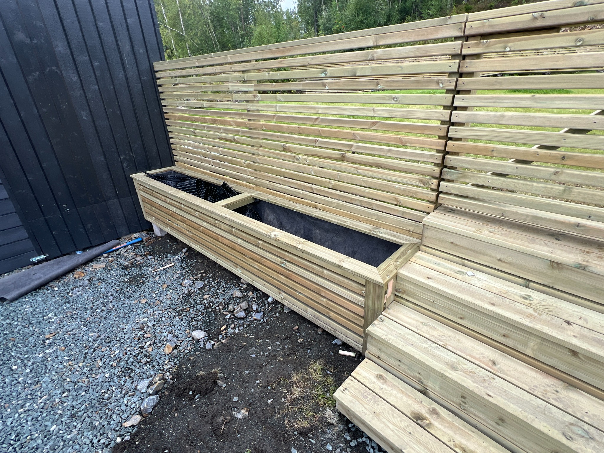 Drewniana konstrukcja z poziomych listew tworząca ściankę i donicę, w tle zielona roślinność, na ziemi szary żwir.