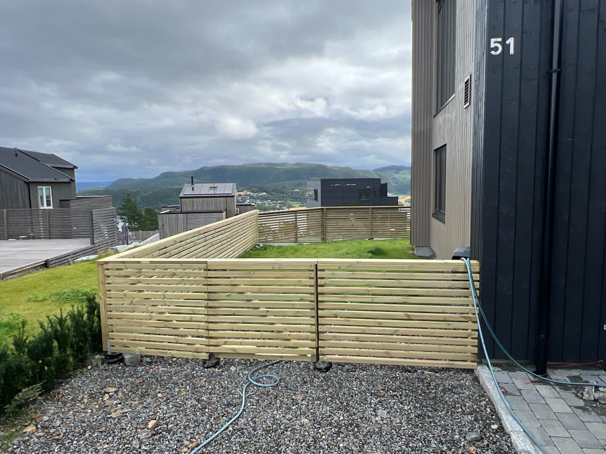 Nowoczesne ogrodzenie panelowe z jasnego drewna na żwirowym podłożu, widok na dom z numerem 51 i górzysty krajobraz w tle.