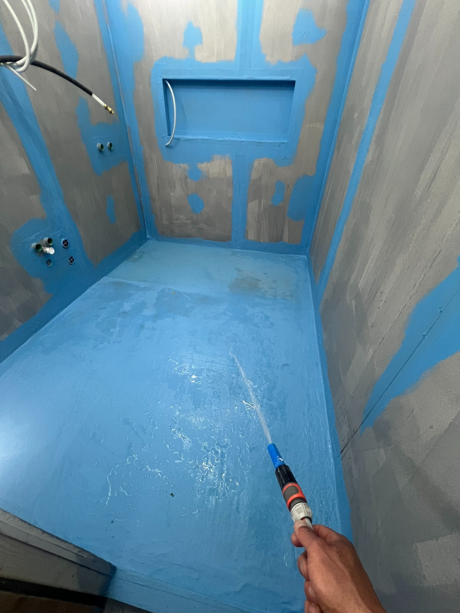 Hydroizolacja w narożnej kabinie prysznicowej z wnęką, ściany pokryte szarą farbą z niebieskimi akcentami, woda spryskiwana z węża w celu sprawdzenia szczelności.
