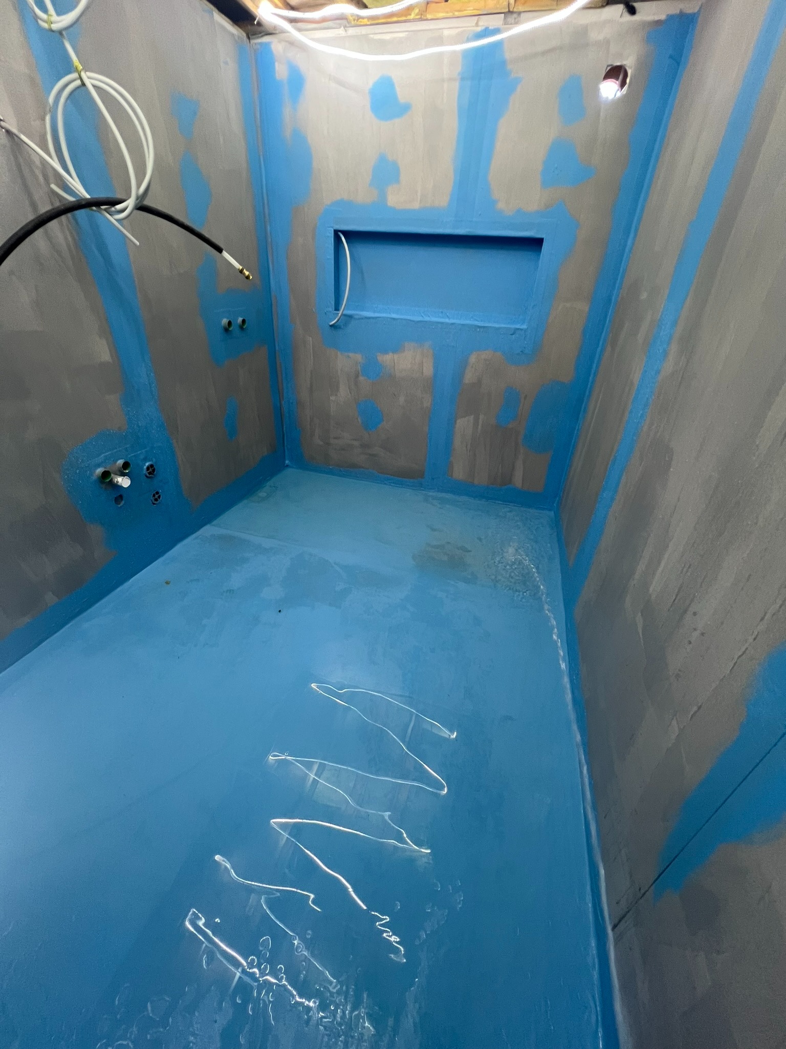 Przygotowanie łazienki do dalszych prac: hydroizolacja w kolorze niebieskim, zabezpieczone taśmą narożniki, widoczne instalacje wodne i elektryczne, wnęka w ścianie.