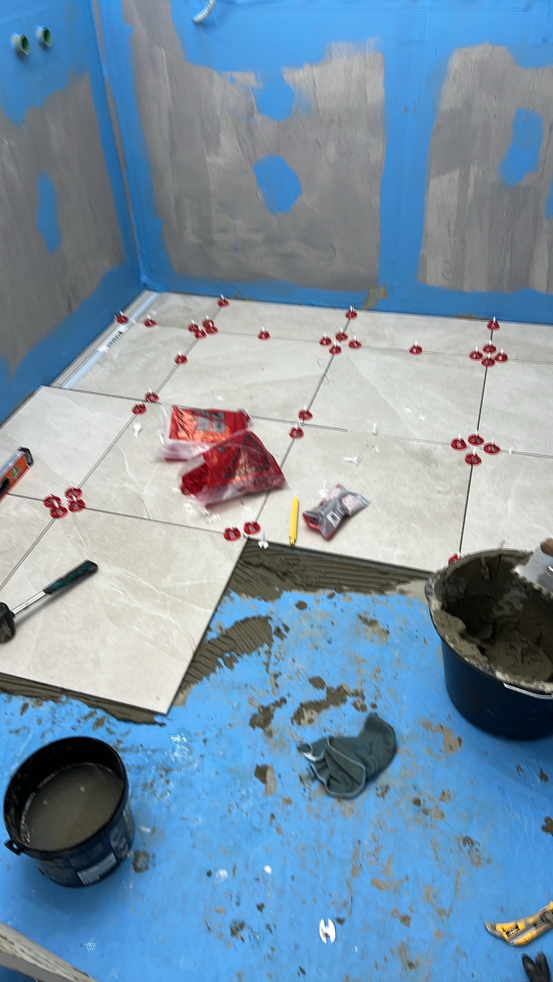 Układanie jasnych płytek podłogowych w łazience z użyciem poziomicy, krzyżyków dystansowych i paczki z zapasem. Na podłodze widoczna warstwa hydroizolacji w kolorze niebieskim oraz wiadro z klejem...