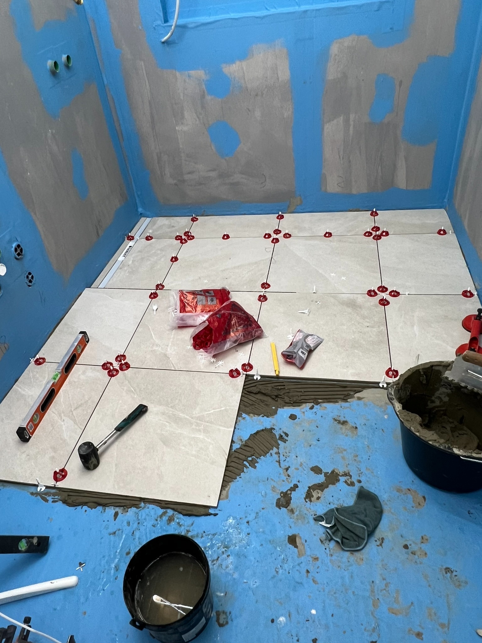 Wykładanie płytek podłogowych w łazience z użyciem poziomicy, gumowego młotka i systemu poziomowania z czerwonymi klipsami, widoczny kubeł z zaprawą i brudna podłoga zabezpieczona folią w kolorze...