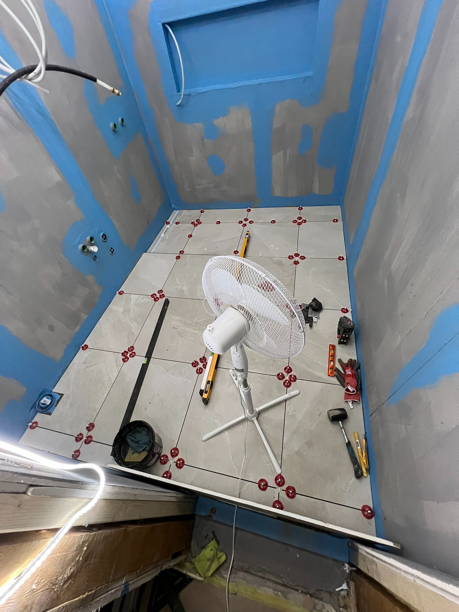 Łazienka w trakcie remontu: położone szare płytki podłogowe z czerwonymi krzyżykami dystansowymi, zabezpieczone ściany i sufit niebieską folią w płynie, wentylator stojący, narzędzia glazurnicze