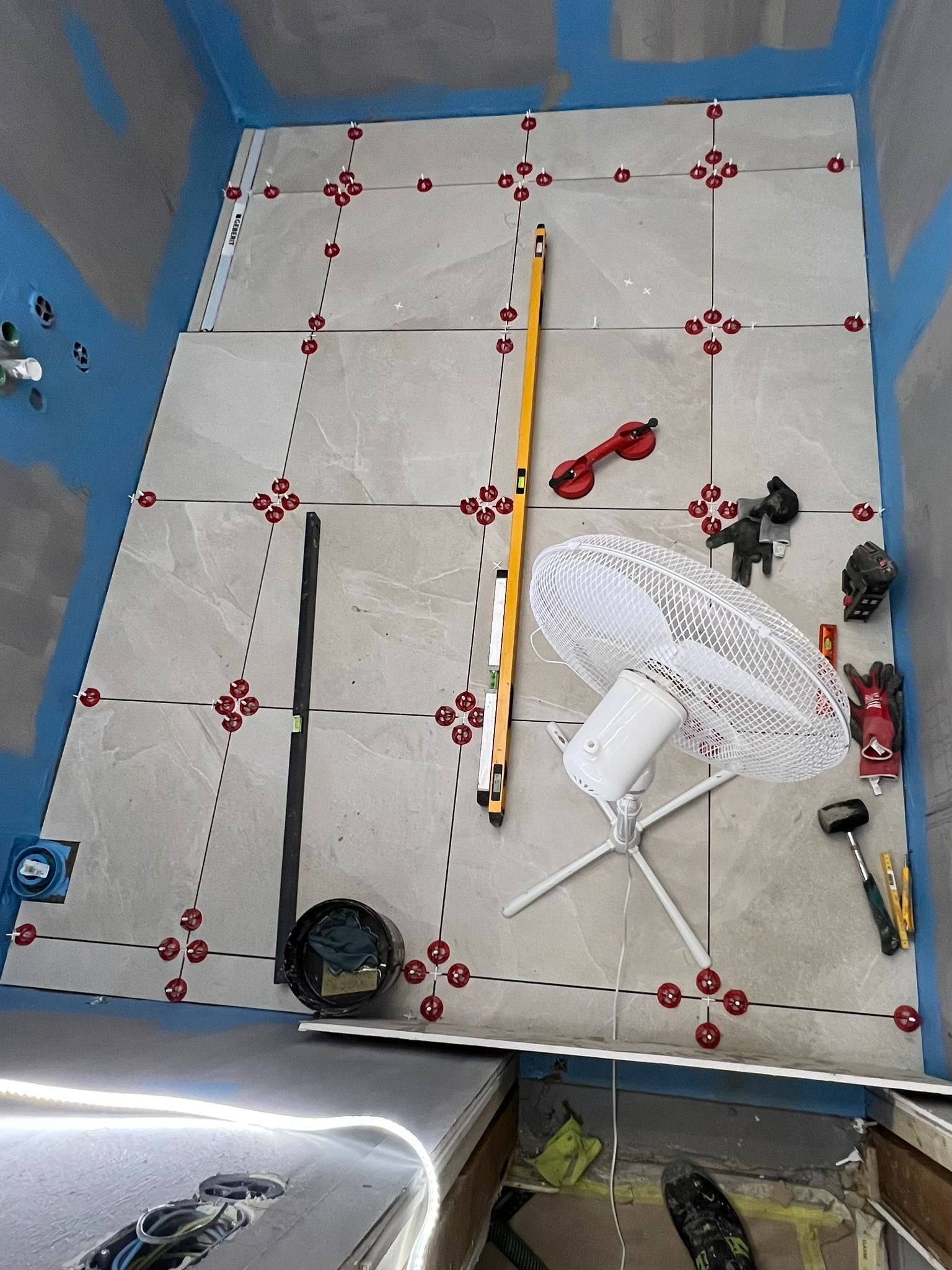 Proces układania płytek podłogowych w łazience z użyciem poziomicy, przyssawki do płytek i systemu poziomowania płytek, widoczny wentylator i narzędzia glazurnicze.