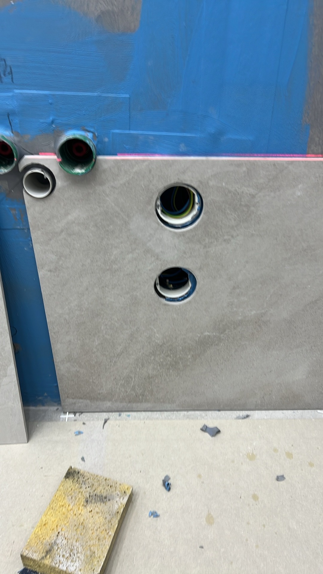 Montaż szarej płyty z wyciętymi otworami na instalacje elektryczne i sanitarne na niebieskiej ścianie, widoczny laserowy poziom, żółta gąbka na podłodze.