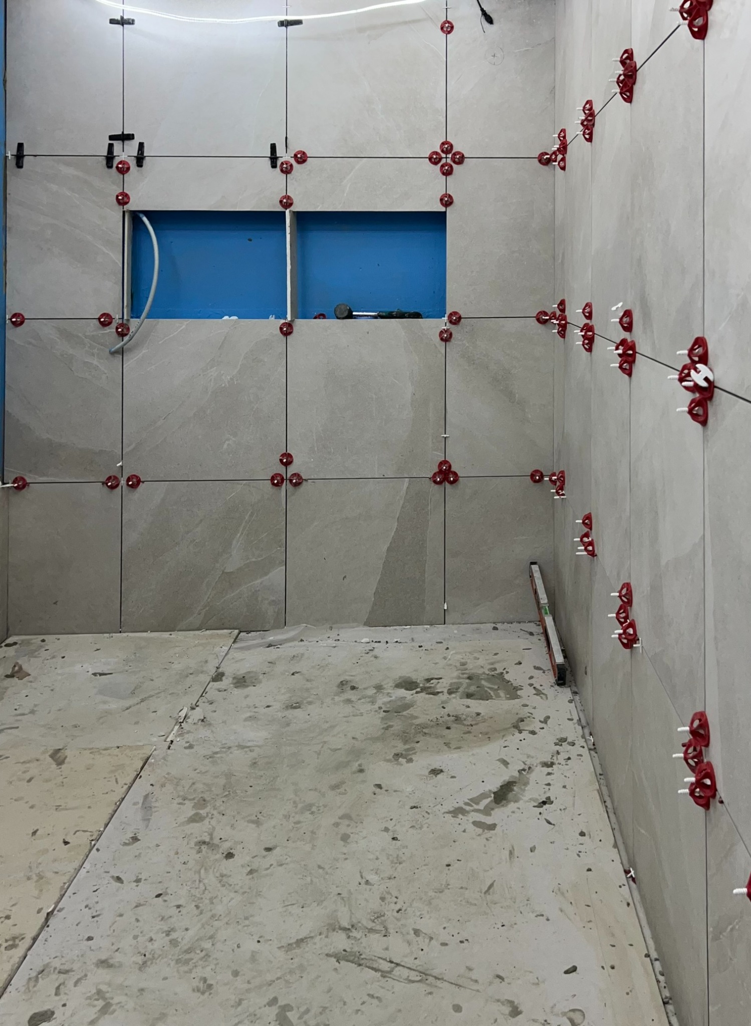 Układanie jasnych, marmurkowych płytek w łazience, z użyciem czerwonych klipsów poziomujących i czarnych krzyżyków dystansowych, widoczny otwór na instalację w ścianie, poziomica przy ścianie.