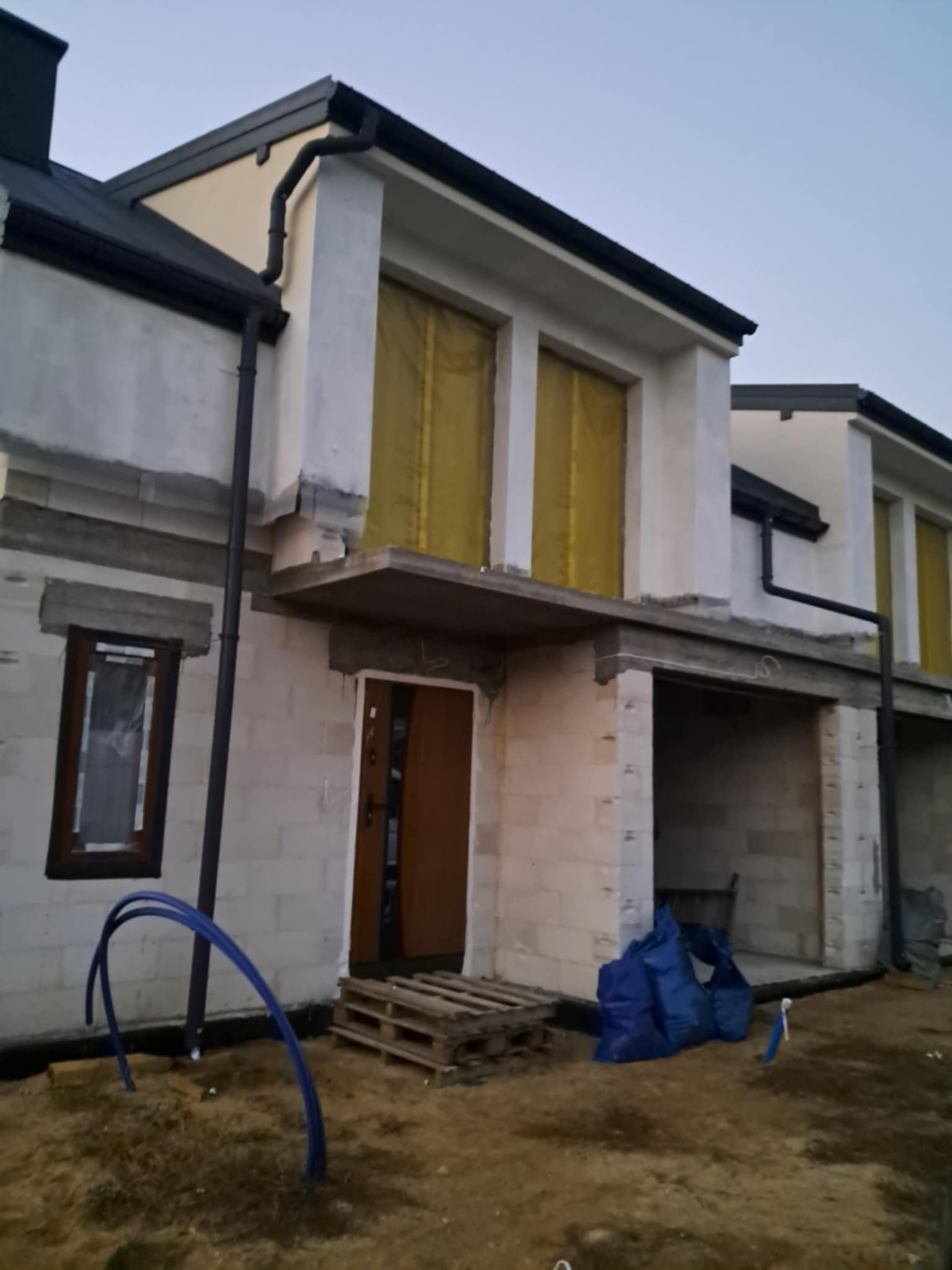 Surowy stan dwukondygnacyjnego domu z pustaków, z widocznymi niezabezpieczonymi oknami, paletą i niebieskimi rurami na placu budowy.