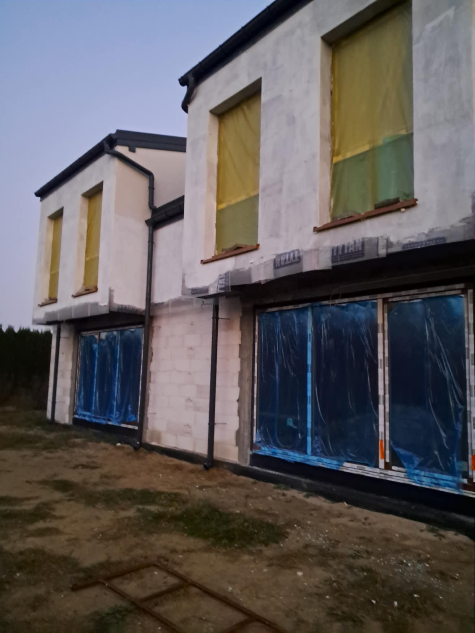 Surowy stan dwukondygnacyjnego domu murowanego z oknami zabezpieczonymi folią, widoczne bloczki betonowe i przygotowane podłoże.