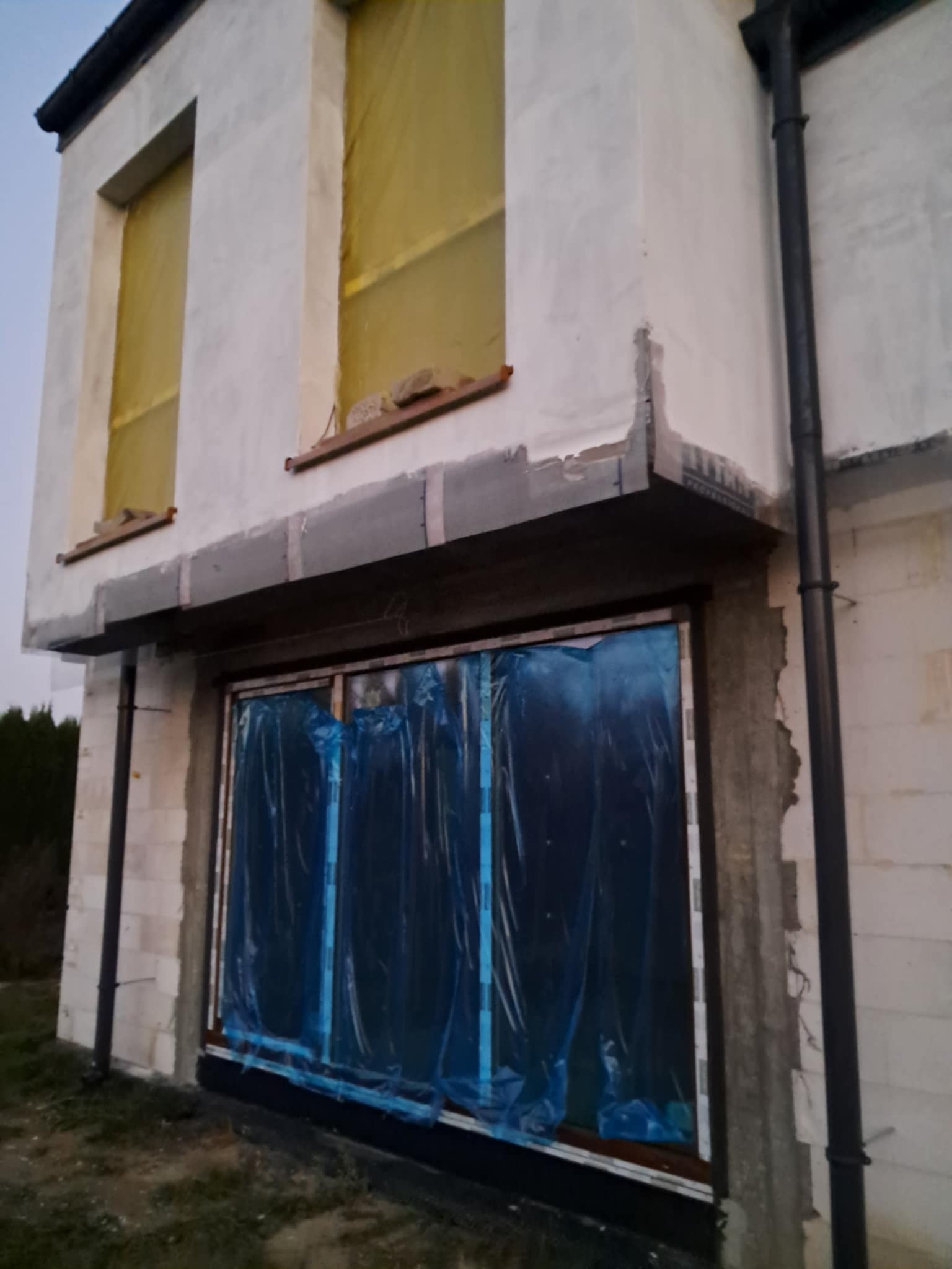 Surowy stan budynku w trakcie budowy, okna zabezpieczone folią, widoczne elementy konstrukcyjne i fragmenty izolacji.