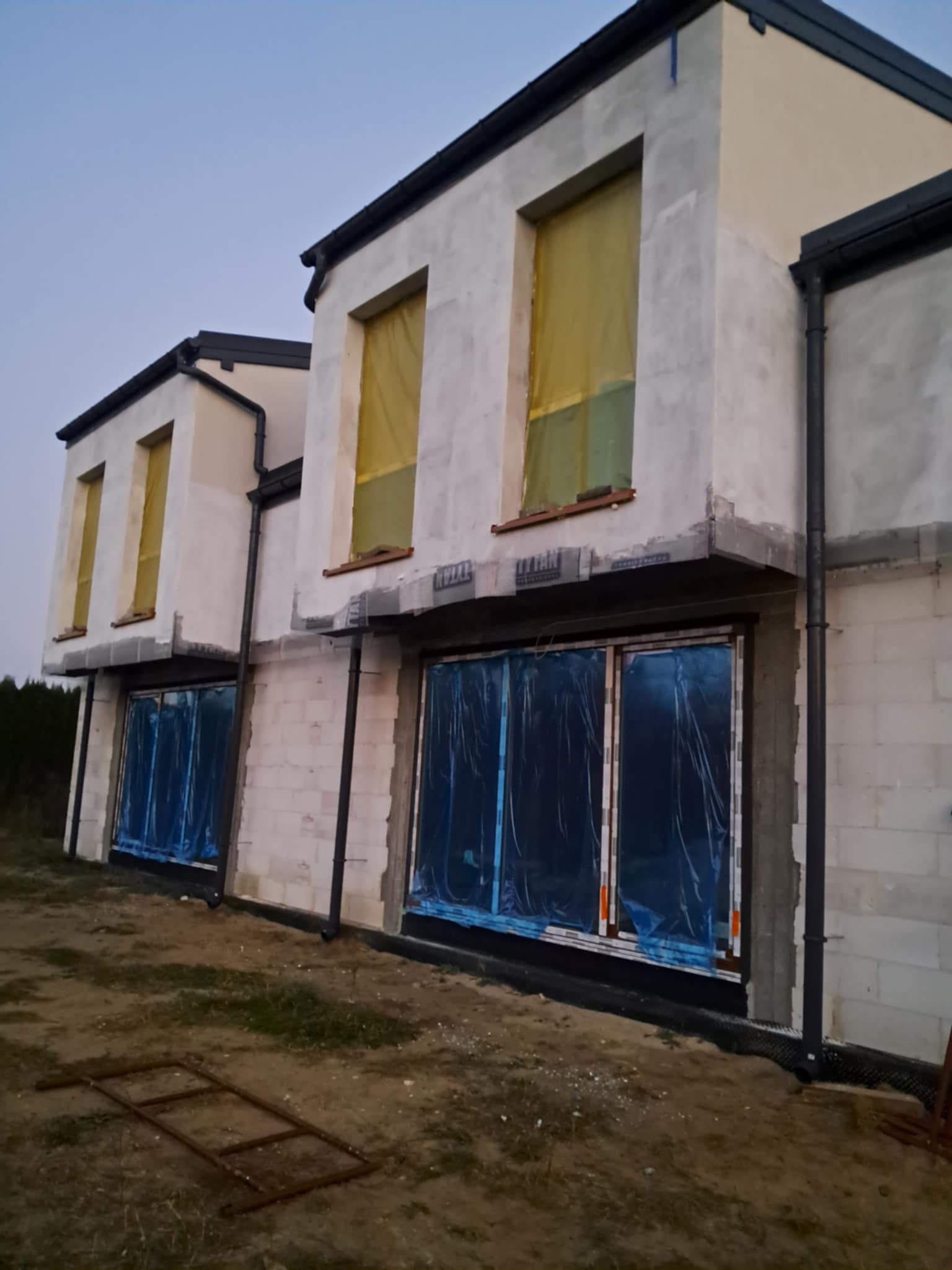 Surowy stan dwukondygnacyjnego domu z pustaków, zabezpieczone okna folią, widoczne rynny i fragment metalowej konstrukcji na trawie.