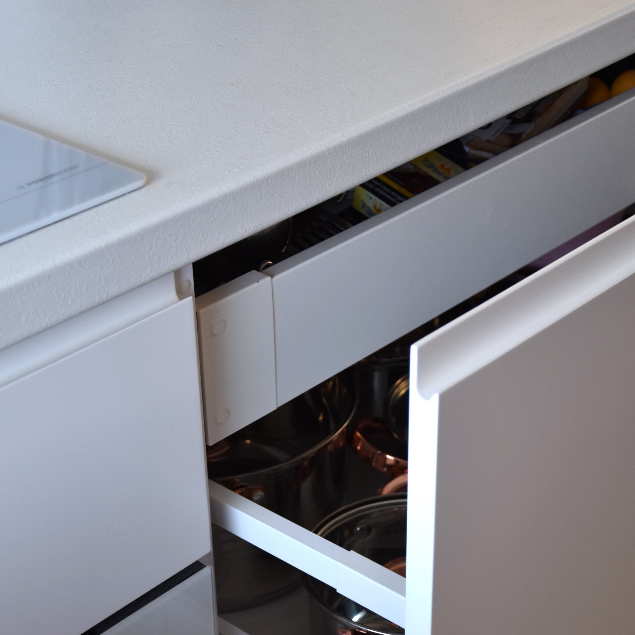 Otwarta szuflada w białej zabudowie kuchennej, wypełniona garnkami z miedzianymi akcentami i różnymi produktami spożywczymi. Widoczny fragment płyty indukcyjnej.