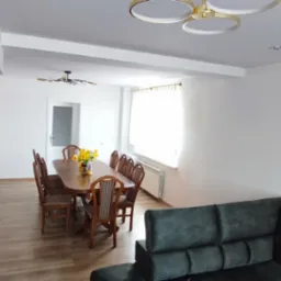 Jasne wnętrze z drewnianą podłogą, stołem i szarym narożnikiem. Ściany białe, oświetlenie sufitowe. Widoczny kominek z okładziną imitującą kamień.