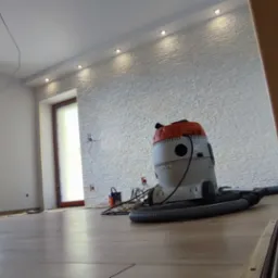 Pokój po remoncie z ułożonymi panelami podłogowymi, widoczny odkurzacz przemysłowy i ściana z dekoracyjną strukturą. Widoczne listwy przypodłogowe i oświetlenie sufitowe.